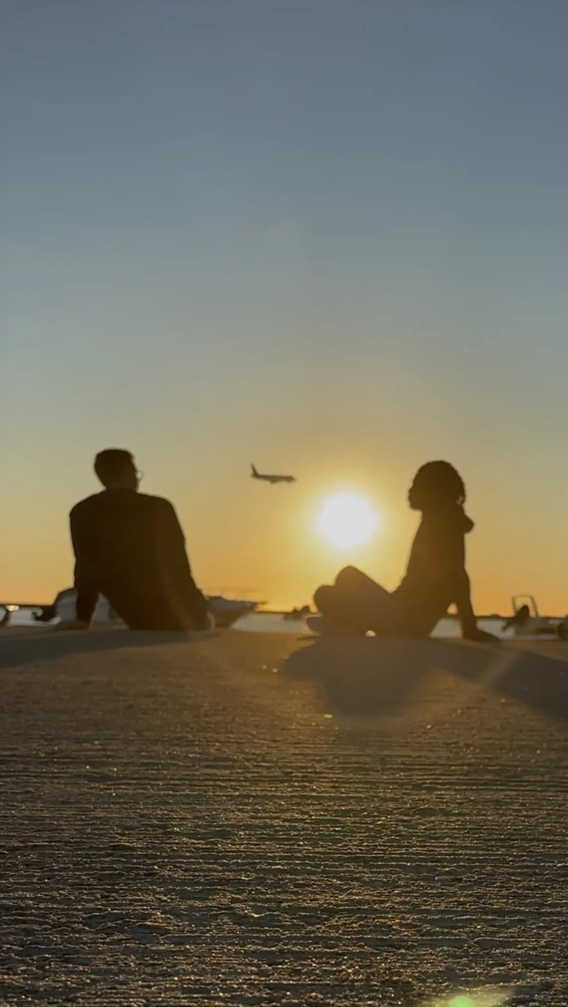 Two people sitting on a beach at sunset while a plane takes off